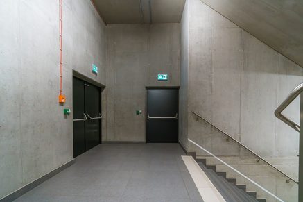 Dark fire doors and illuminated exit signs beside a concrete stairwell with a metal handrail